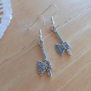May include: Silver axe earrings with detailed blade and handle design. The earrings have a hook closure and are displayed on a light wooden surface. The axes are a miniature size, perfect for a unique accessory.