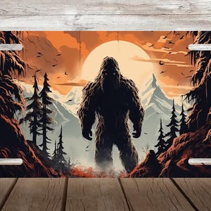 May include: License plate with a Bigfoot illustration. The artwork shows the creature standing before a sunset, with mountains and trees in the background. The colour scheme includes shades of orange, brown, and green.