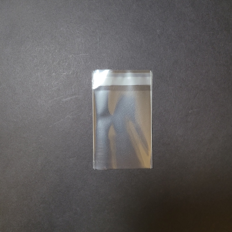 May include: A clear plastic sleeve with a white adhesive strip at the top. The sleeve is designed to protect and store items.