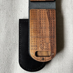 May include: A wooden cribbage board with a dark finish, featuring numerous drilled holes for peg placement. The board is encased in a black leather sleeve. The design includes a curved section and a metal snap closure.