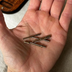 May include: A collection of small, metallic, cylindrical pins with varying designs. Some pins have grooved ends, while others have a smooth, rounded appearance. The pins are a warm, brassy color and appear to be made of metal.