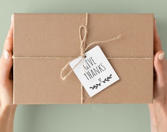 May include: A rectangular brown gift box tied with twine and a white tag that reads "GIVE THANKS" in black lettering. The tag is decorated with a small heart and leaf design. The box is held by two hands.