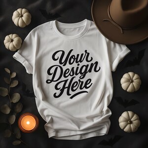 May include: A white t-shirt with the text "Your Design Here" in a black script font. The shirt is surrounded by small white pumpkins, a brown hat, a lit candle, and decorative leaves, all set against a dark background with bat silhouettes.