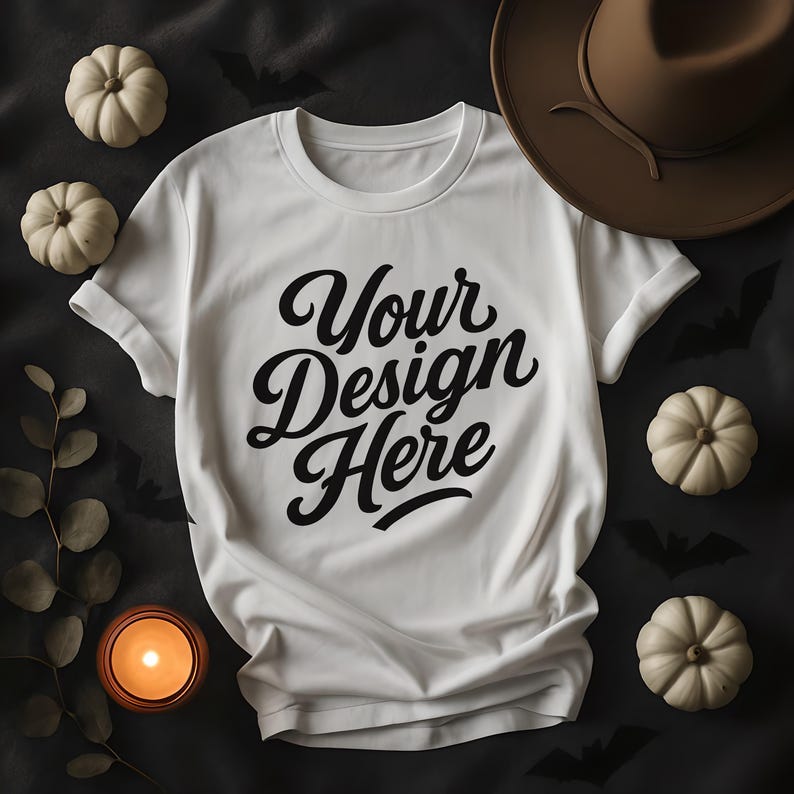 May include: A white t-shirt with the text "Your Design Here" in a black script font. The shirt is surrounded by small white pumpkins, a brown hat, a lit candle, and decorative leaves, all set against a dark background with bat silhouettes.