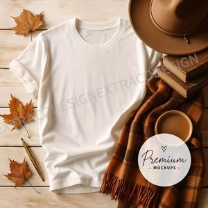 Flat lay of a blank oversized white t-shirt on a wooden surface, styled with a plaid scarf, warm chai tea, dried maple leaves, and vintage books for a cozy autumn aesthetic.