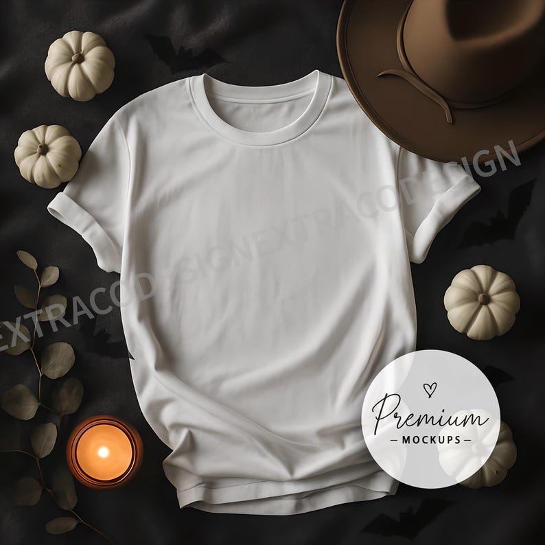 Flat lay of an oversized white t-shirt on a soft charcoal gray fleece blanket. Surrounding the shirt are Halloween elements: mini white pumpkins, a few scattered black bats cut from paper, a lit candle in a glass jar, and dried eucalyptus leaves. The mood is moody yet warm, lit with soft ambient lighting from the candle and side daylight. The white shirt is centered and blank, ready for overlay designs. Square format, styled for a cozy yet elevated Halloween aesthetic.