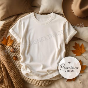 Oversized white t-shirt laid flat on a chunky beige knit throw, surrounded by neutral pillows, scattered fall leaves, and soft golden light for a calm, seasonal vibe.
