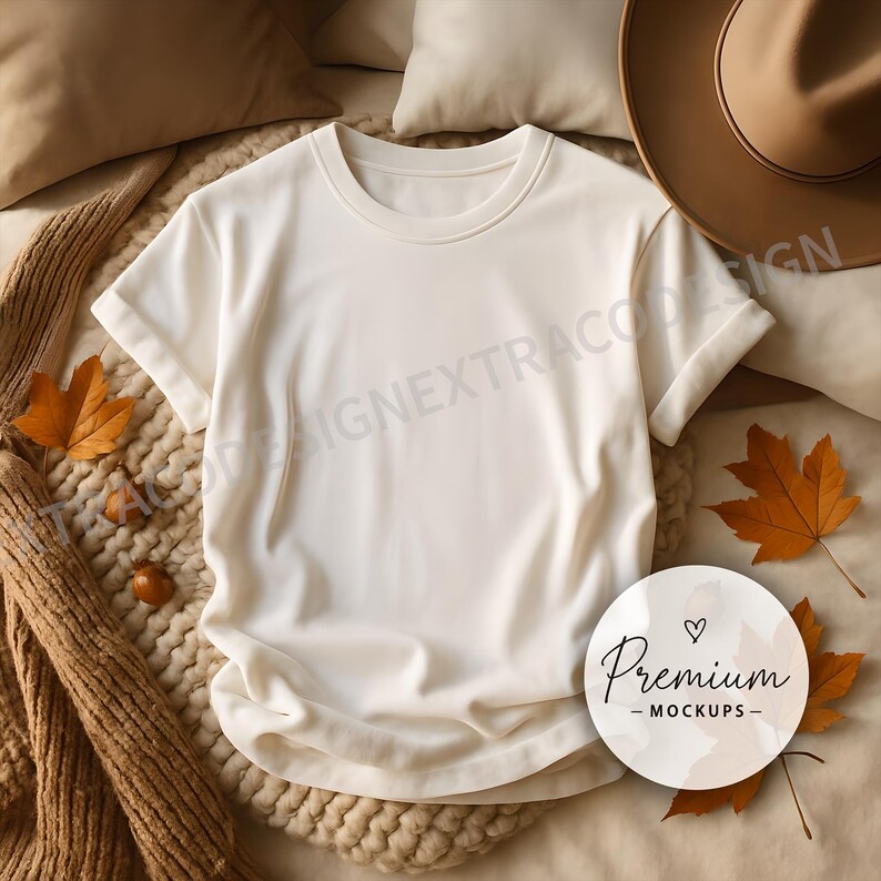 Oversized white t-shirt laid flat on a chunky beige knit throw, surrounded by neutral pillows, scattered fall leaves, and soft golden light for a calm, seasonal vibe.