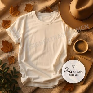 White t-shirt mockup on a textured blanket with cinnamon sticks, a ceramic mug, and dried autumn leaves styled in warm, natural tones with soft side lighting.