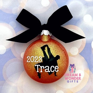 May include: A red and gold glitter Christmas ornament with a black silhouette of a person doing a gymnastics pose. The ornament has the year 2023 and the name "Trace" written on it.