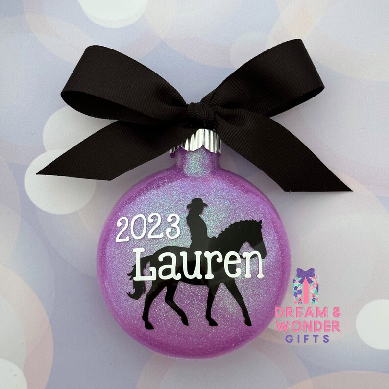 May include: Purple glitter Christmas ornament with a black silhouette of a person riding a horse. The ornament is personalized with the name "Lauren" and the year "2023".