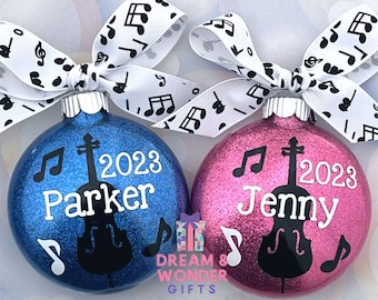 May include: Two personalized Christmas ornaments with glitter finish. One is blue with the name "Parker" and the year "2023" and the other is pink with the name "Jenny" and the year "2023". Both ornaments feature a black silhouette of a cello and musical notes.
