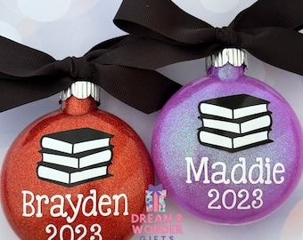 May include: Two personalized Christmas ornaments with a stack of books design. One ornament is red glitter with the name Brayden and the year 2023. The other ornament is purple glitter with the name Maddie and the year 2023.