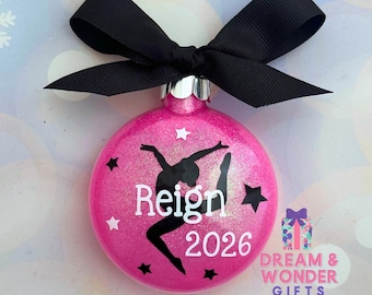 Op de afbeelding: Een ronde, glitterende roze kerstbal met een zwarte silhouet van een turner in een spronghouding. De naam Reign en het jaartal 2026 zijn in witte letters gedrukt. De kerstbal is afgewerkt met een grote, zwarte strik van grosgrain lint.