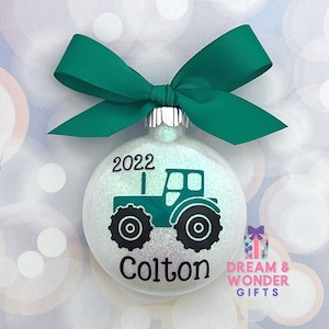 May include: A white glitter Christmas ornament with a green tractor design, the year 2022, and the name Colton. The ornament has a green ribbon.