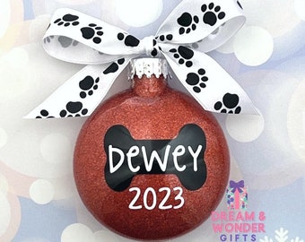 May include: A red glitter Christmas ornament with a black bone shape and the name "Dewey" and the year "2023" written in white. The ornament is tied with a white ribbon with black paw prints.