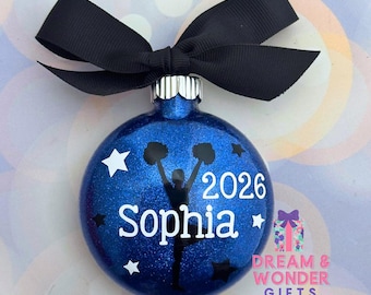 May include: A round, blue glitter Christmas ornament topped with a black ribbon bow. The ornament features a black silhouette of a cheerleader with pom-poms, the white text "Sophia" and "2026", and several white and black star shapes.