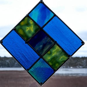 Op de afbeelding: Een ruitvormig glas-in-loodraam met blauwe en groene glasstukken. Het raam is omlijst met zwart metaal.