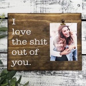 May include: A brown wooden picture frame with a white inscription that reads "I love the shit out of you." A photo of a couple is clipped to the frame with a gold clip.
