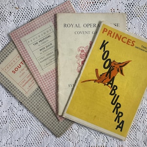 May include: A selection of old theatre programmes and playbills. Titles include "Kookaburra" and "The Proposal." The covers are in various colours, including yellow, red, and white, with text and illustrations. The items are arranged on a white lace surface.