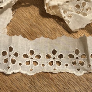 May include: Close-up of off-white eyelet lace trim with a scalloped edge. The trim features a floral design with openwork details. The fabric is layered, showing the texture and delicate nature of the material. Ideal for sewing and craft projects.