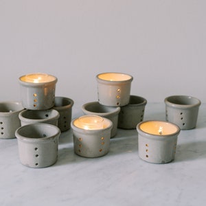 May include: A group of gray ceramic candle holders with small holes, some with lit tea lights inside. The holders are arranged in a pyramid shape on a white marble surface.