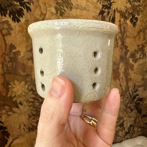May include: A small, light gray ceramic container with several evenly spaced holes. The container has a slightly textured surface and a rounded rim. The object is held in a hand, with a patterned background.