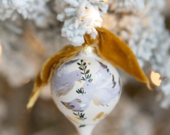 May include: A white Christmas ornament with a purple and gold design of two birds and foliage. The ornament is hanging from a gold velvet ribbon on a snowy Christmas tree.