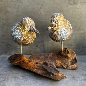 Peut inclure: Deux figurines d'oiseaux ronds avec une finition texturée et craquelée dans des tons dorés, bruns et bleus. Chaque oiseau est monté sur une fine tige métallique fixée à un morceau de bois flotté brun foncé, formant une sculpture décorative.