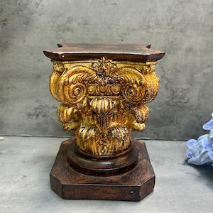 May include: An antique-style pedestal with a square base and an ornate, gold-coloured column. The column has intricate carvings and a dark brown top. The pedestal is likely made of wood and is suitable for displaying small items.