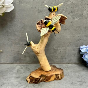 May include: A wooden sculpture of a tree branch with three decorative bees. The bees are black and yellow with white wings. The sculpture is on a wooden base and is surrounded by flowers.
