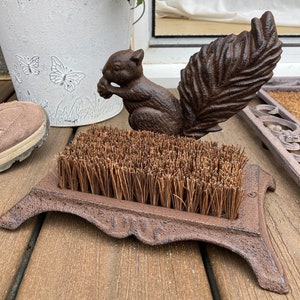 May include: A cast iron boot scraper with a squirrel design. The scraper has a brown finish and a stiff brush made of natural fibers.