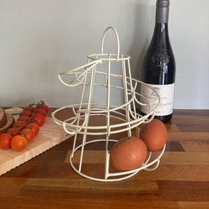 May include: Cream-coloured, spiral wire egg holder containing two brown eggs. A dark wine bottle and a cluster of red cherry tomatoes on a wooden board are also visible. The egg holder has a handle for carrying.