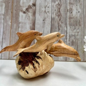 Peut inclure: Une sculpture en bois repr&eacute;sentant trois dauphins sautant d'une base en loupe de bois. Les dauphins sont sculpt&eacute;s dans du bois clair, mettant en valeur leurs formes profil&eacute;es. La base a un aspect naturel textur&eacute;.
