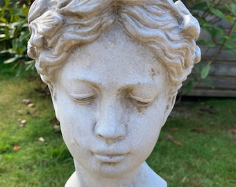 May include: A weathered, gray stone bust of a serene face with closed eyes and detailed curly hair. The sculpture is mounted on a textured pedestal, set against a backdrop of green grass and foliage. A classical garden statue.