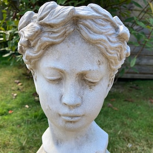 May include: A weathered, grey stone bust of a serene face with closed eyes and detailed curly hair. The sculpture is mounted on a textured pedestal, set against a backdrop of green grass and foliage. A classical garden statue.