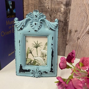 May include: A small, blue, distressed picture frame with a decorative design. The frame is standing on a white surface with pink flowers.