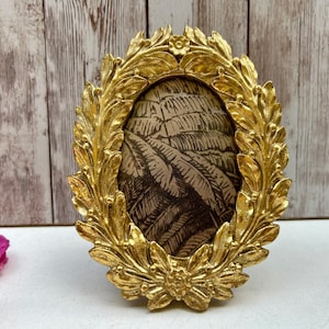 May include: Oval gold picture frame with a detailed leaf and floral design. The frame holds a sepia-toned print. The frame is approximately 15 cm tall and 10 cm wide, with a decorative border.