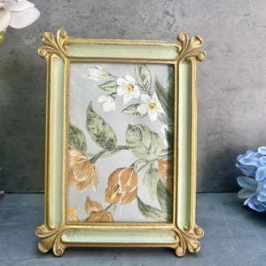 May include: A rectangular picture frame with a pale green border and gold detailing. The frame contains a floral print with white and orange flowers and green leaves. The frame is standing upright.