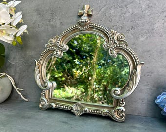 May include: Ornate silver-colored mirror with a crown-like design. The mirror has a decorative frame with floral and scroll details. The mirror reflects a green, natural outdoor scene. A decorative piece for home decor.