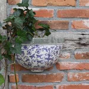 Peut inclure: Un pot en céramique blanc avec un motif floral bleu et blanc, suspendu à un mur de briques. Le pot est rempli de lierre vert.