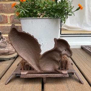May include: A brown cast iron squirrel boot scraper with a brush underneath. The scraper is on a wooden deck.