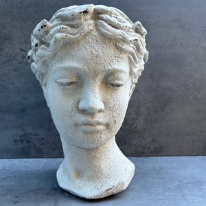 May include: A distressed, off-white sculpture of a woman's head with closed eyes. The classical bust features detailed facial features and a textured surface, suggesting an aged appearance. The head is adorned with a detailed hairstyle.
