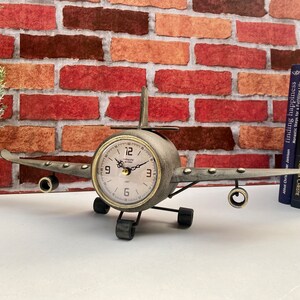May include: A metal aeroplane clock with a brown and silver finish. The clock face is white with black numbers and hands. The aeroplane has a propeller and wings.