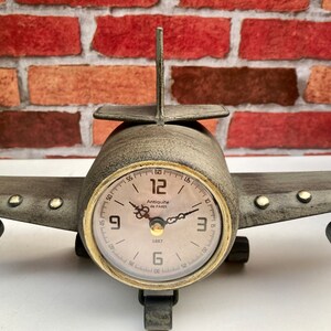 May include: A vintage airplane-shaped clock with a brown metal body and a cream-coloured face. The clock face has black Roman numeral hour markers and black hands. The clock reads "Antique de Paris 1887".