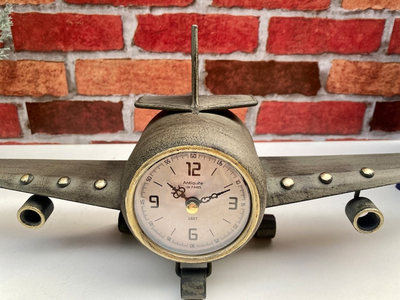 May include: A vintage airplane-shaped clock with a brown metal body and a cream-coloured face. The clock face has black Roman numeral hour markers and black hands. The clock reads "Antique de Paris 1887".