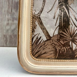 May include: A white picture frame with a gold trim and a brown and white tropical print inside. The frame has a beaded design around the edge.