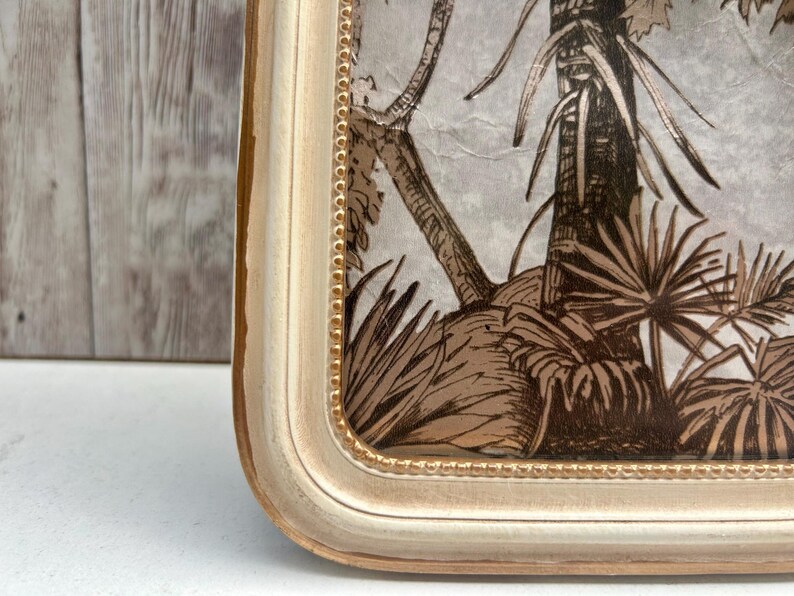 May include: A white picture frame with a gold trim and a brown and white tropical print inside. The frame has a beaded design around the edge.