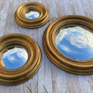 May include: Three gold-framed round mirrors with a reflective surface. The mirrors are nested inside each other, with the largest mirror in the back and the smallest mirror in the front. The mirrors reflect a cloudy sky.