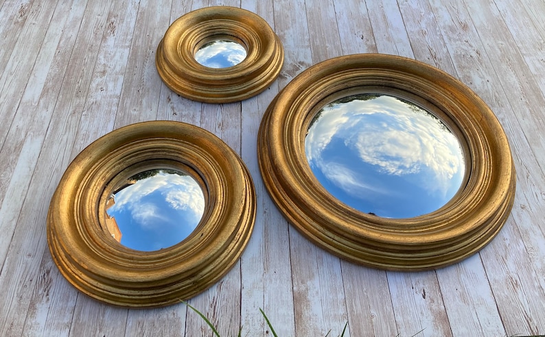 May include: Three gold-framed round mirrors with a reflective surface. The mirrors are nested inside each other, with the largest mirror in the back and the smallest mirror in the front. The mirrors reflect a cloudy sky.